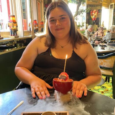 A medium shot of Frankie, a woman with brown hair, sitting at a table in a brightly decorated restaurant. She is wearing a black sleeveless dress and smiling at the camera. In front of her is a small red bowl containing a dessert with a single lit candle, which is resting on a base that is emitting a thick cloud of white dry ice smoke across the table.