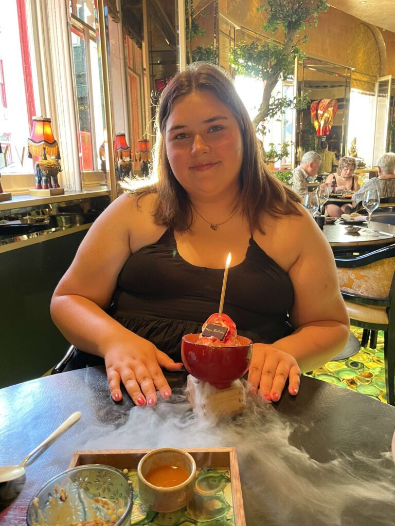 A medium shot of Frankie, a woman with brown hair, sitting at a table in a brightly decorated restaurant. She is wearing a black sleeveless dress and smiling at the camera. In front of her is a small red bowl containing a dessert with a single lit candle, which is resting on a base that is emitting a thick cloud of white dry ice smoke across the table.