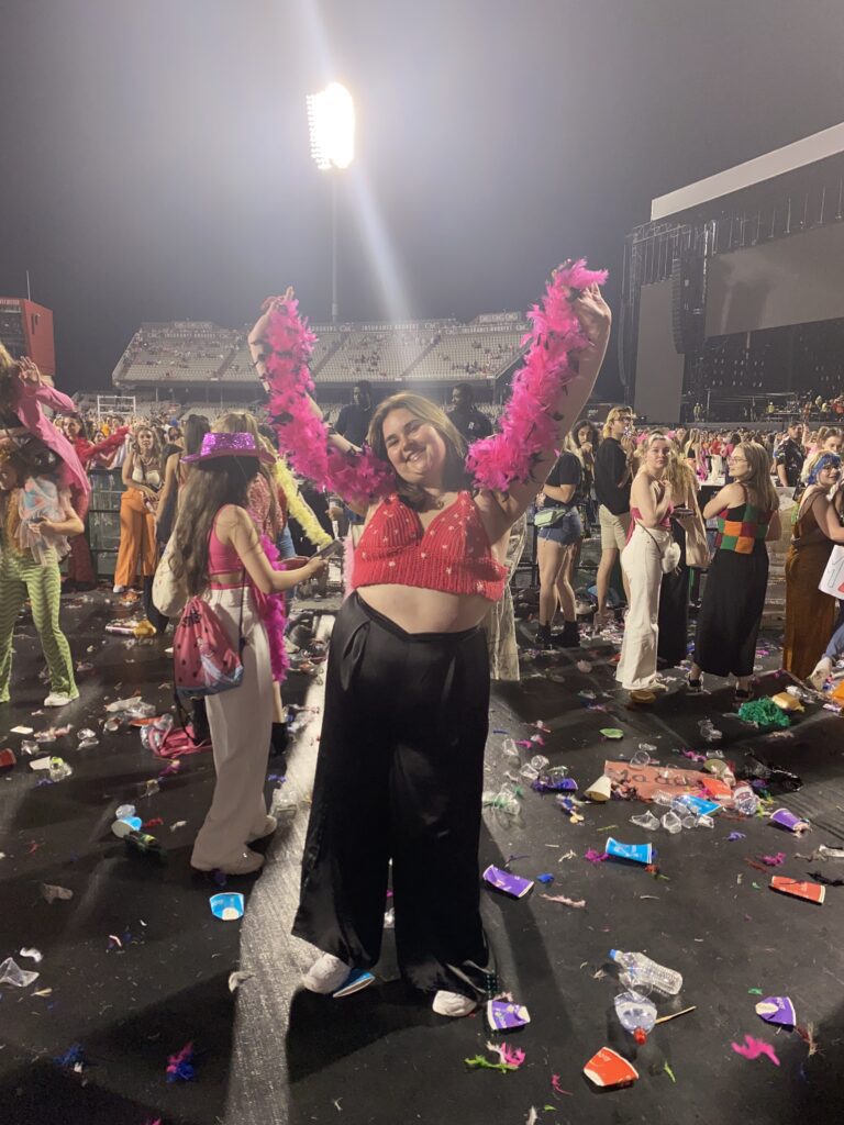 A full-length photo of Frankie smiling with her arms raised at an outdoor concert at night. She is wearing a red, star-patterned crochet crop top, black wide-leg trousers, and a bright pink feather boa draped over her arms. The ground around her is covered in colourful confetti and discarded cups, and the illuminated stadium stands and concert stage are visible in the background.