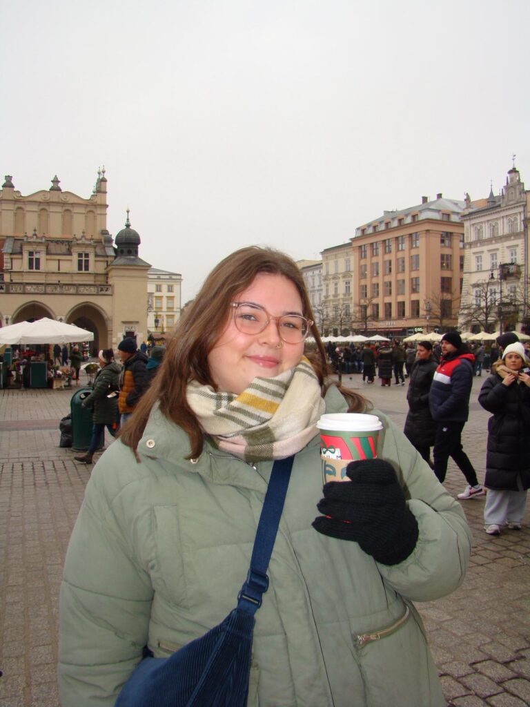 A medium shot of Frankie, a woman with brown hair and clear-framed glasses, standing in a cobblestone square in Krakow. She is wearing a sage green puffer jacket, a plaid scarf, and black gloves, while holding a red and green Starbucks holiday-themed coffee cup. Behind her are historic buildings, including a large arched structure, under an overcast sky.