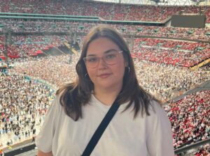 Frankie stands at a railing overlooking a sea of fans at an Oasis concert. She is wearing a simple white t-shirt, black cross-body bag and round glasses, looking happy as she poses against the backdrop of a sunlit stadium.