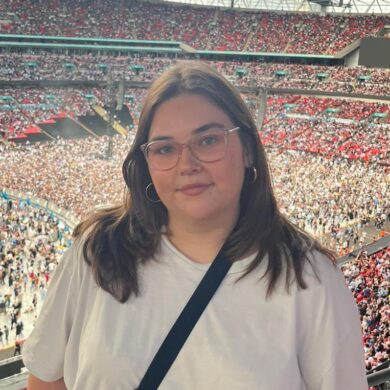 Frankie stands at a railing overlooking a sea of fans at an Oasis concert. She is wearing a simple white t-shirt, black cross-body bag and round glasses, looking happy as she poses against the backdrop of a sunlit stadium.