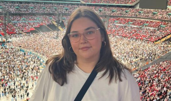Frankie stands at a railing overlooking a sea of fans at an Oasis concert. She is wearing a simple white t-shirt, black cross-body bag and round glasses, looking happy as she poses against the backdrop of a sunlit stadium.