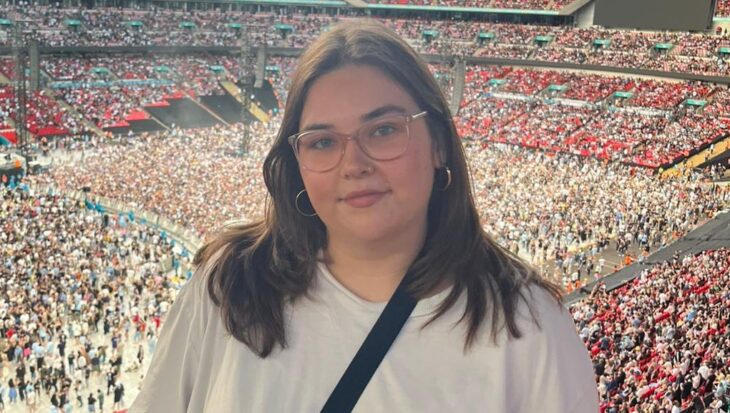 Frankie stands at a railing overlooking a sea of fans at an Oasis concert. She is wearing a simple white t-shirt, black cross-body bag and round glasses, looking happy as she poses against the backdrop of a sunlit stadium.