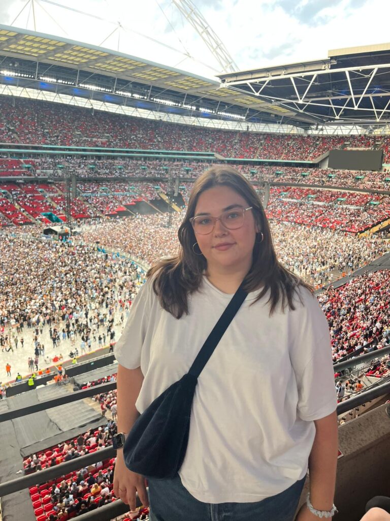 Frankie stands at a railing overlooking a sea of fans at an Oasis concert. She is wearing a simple white t-shirt, black cross-body bag and round glasses, looking happy as she poses against the backdrop of a sunlit stadium.