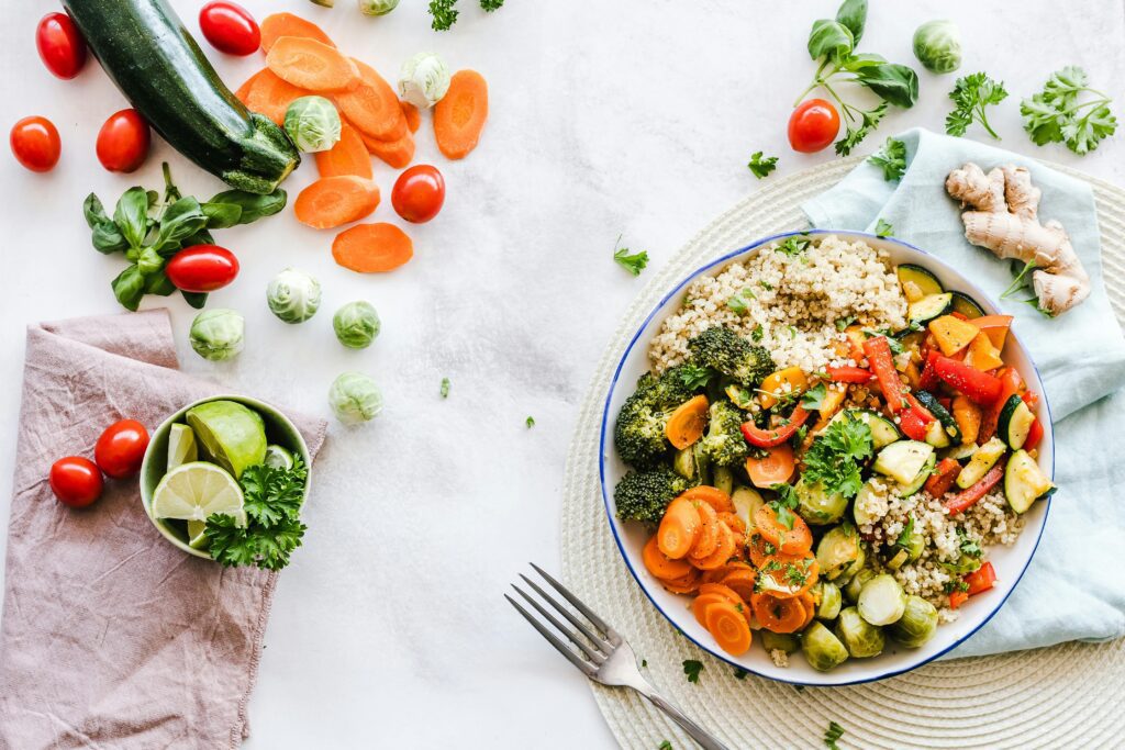 An overhead flat lay shows a colorful, healthy, and balanced meal composed primarily of fresh vegetables. On the right, a white and blue patterned bowl is filled with cooked quinoa, broccoli florets, sliced carrots, brussels sprouts, and sautéed red and yellow bell peppers and zucchini. A fork rests below the bowl. On the left, fresh ingredients are scattered across a white surface, including a whole zucchini, sliced carrots, cherry tomatoes, and scattered brussels sprouts. A small bowl holds lime slices and parsley, next to a folded beige napkin. A piece of ginger root is also visible near the top right of the composition.