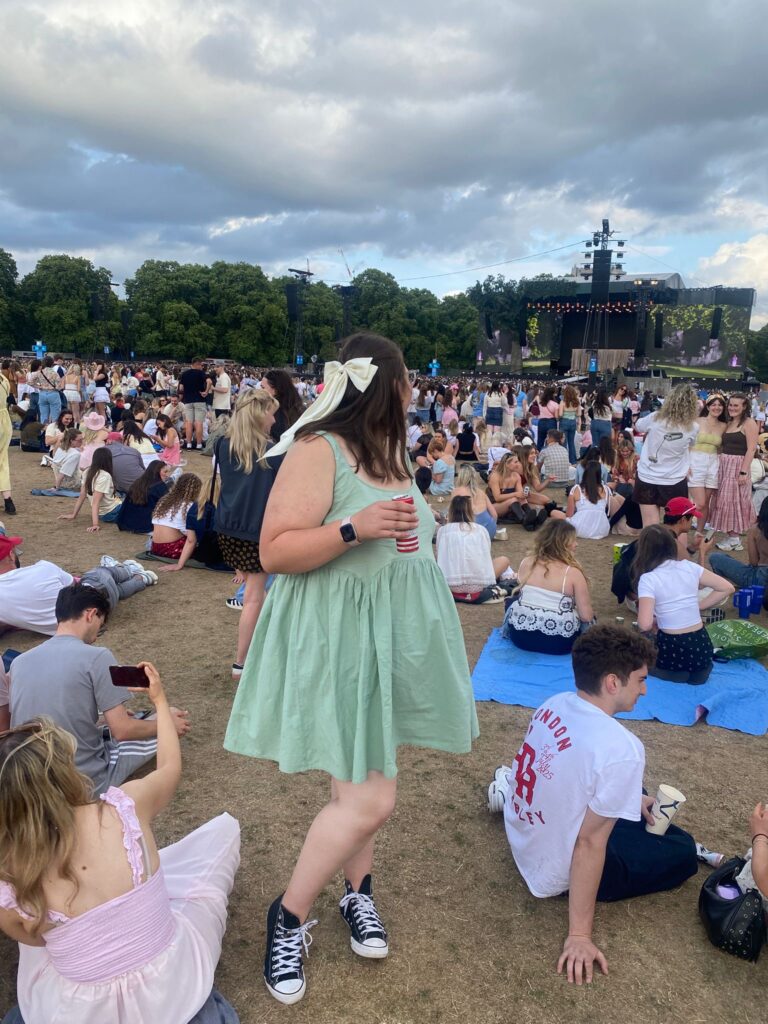 Frankie enjoys the atmosphere at a Sabrina Carpenter concert. She is dressed in a summer-style green dress and a bow, standing amidst a crowd of fans seated on the ground. In the distance, a large stage with video screens is set against a backdrop of green trees and a cloudy sky.