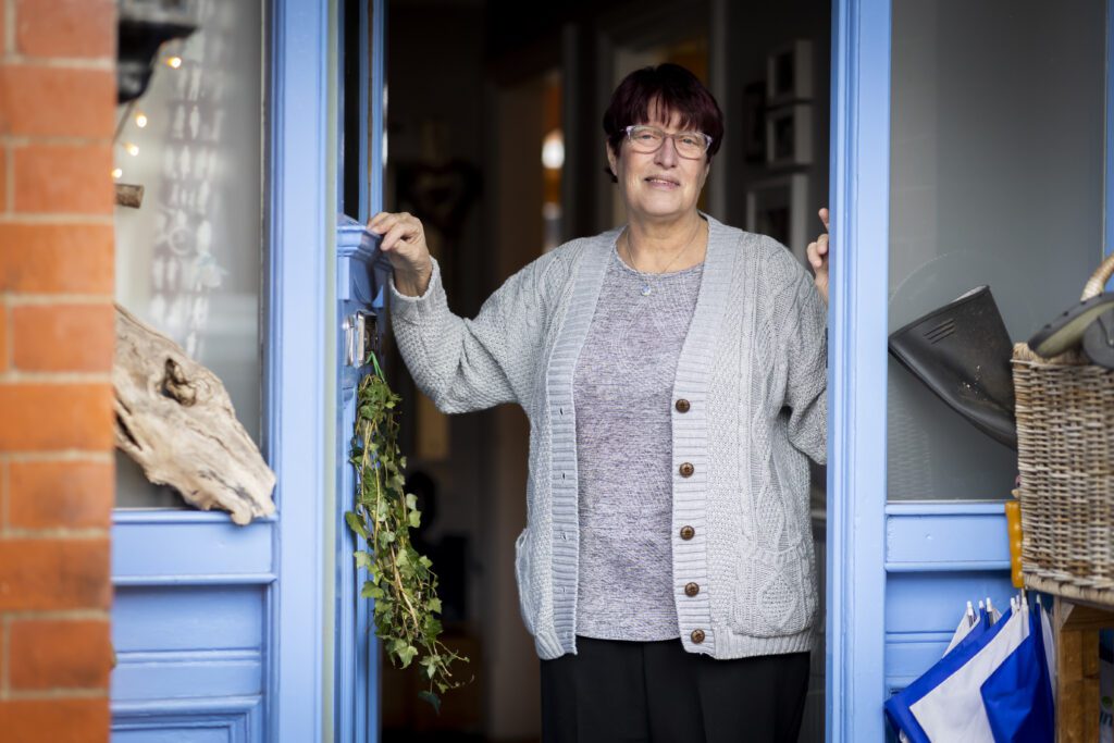 A medium shot shows a smiling woman with short, dark red hair and glasses standing in the doorway of her home. She is wearing a grey marl t-shirt under a light grey button-down cardigan and black trousers. Her right hand rests on the blue door frame, and her left hand is visible on the other side. The doorway is light blue, and to the left of the door is a brick wall. A bundle of green ivy hangs from the door frame near her right hand. To the right of the doorway, a wicker basket and a black boot are visible. The background inside the house is dark and slightly out of focus.