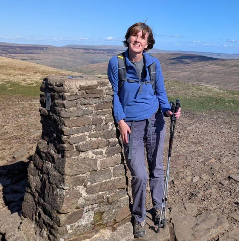 A full-length photo shows Fiona standing at the summit of Pen-y-ghent. She is smiling and leaning against a stone trig point built from stacked rocks. Fiona is wearing a blue long-sleeved athletic top, grey hiking trousers, a backpack, and is holding trekking poles. The background reveals a vast, rolling landscape of brown and green hills under a bright, clear blue sky.