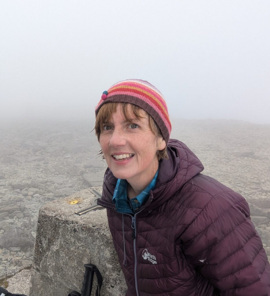 A medium shot shows Fiona at the summit of Ben Nevis. She is a woman with short brown hair, wearing a striped pink and purple beanie and a dark purple North Ridge puffer jacket over a blue plaid shirt. She is smiling and leaning against a concrete trig point on the rocky, fog-covered mountain peak. The background is a dense, white mist that obscures any view beyond the immediate rocky ground.
