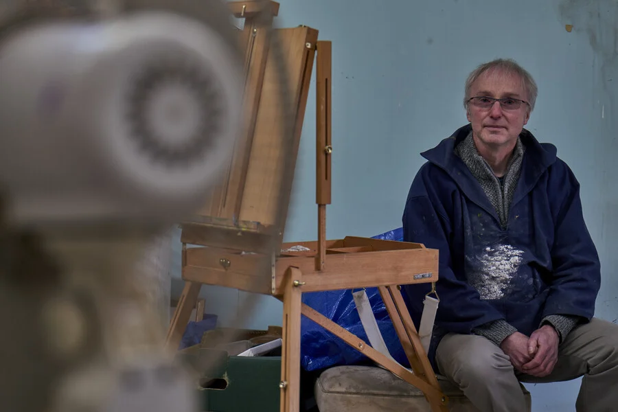  A medium shot shows a man with graying hair and glasses sitting in an artist's studio. He wears a blue smock stained with white paint over a gray sweater and khaki pants. He sits next to a wooden French easel that is set up but currently empty of a canvas. In the blurred foreground, a large, out-of-focus white object partially obscures the left side of the frame. The background consists of a plain, light blue wall with some minor scuffs and marks.