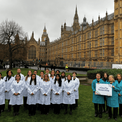 A group of approximately twenty people stand on a grassy lawn in front of the Palace of Westminster in London. Most of the group are wearing white lab coats, while four people on the right wear teal lab coats and hold a sign that reads 