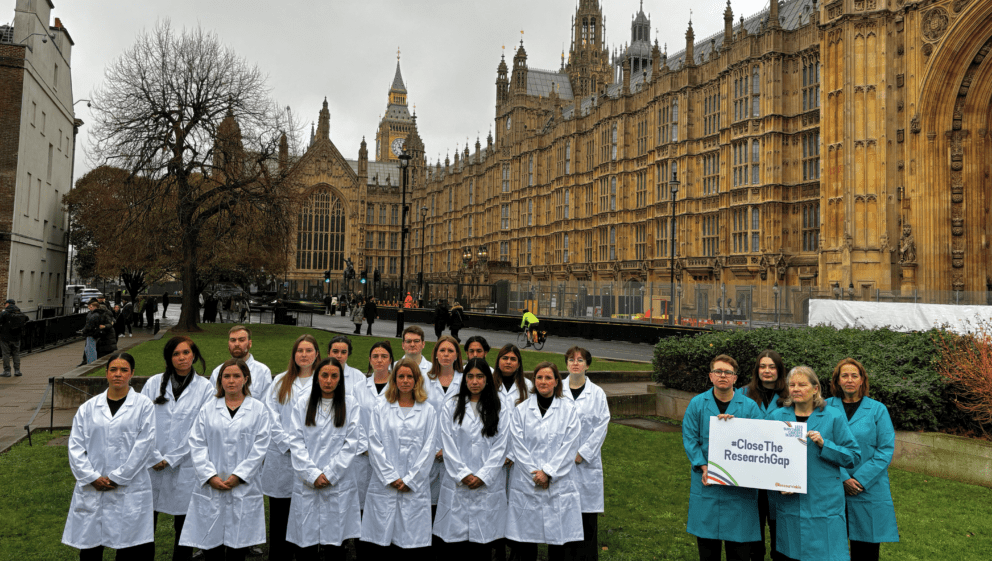A group of approximately twenty people stand on a grassy lawn in front of the Palace of Westminster in London. Most of the group are wearing white lab coats, while four people on the right wear teal lab coats and hold a sign that reads "#CloseTheResearchGap." The Elizabeth Tower, featuring Big Ben, is visible in the background under an overcast sky.