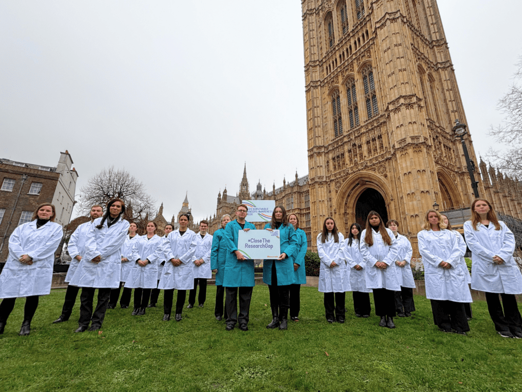 A group of approximately twenty people stand on a grassy lawn in front of the Palace of Westminster in London. Most of the group are wearing white lab coats, while four people in the centre wear teal lab coats and hold a sign that reads "#CloseTheResearchGap." 