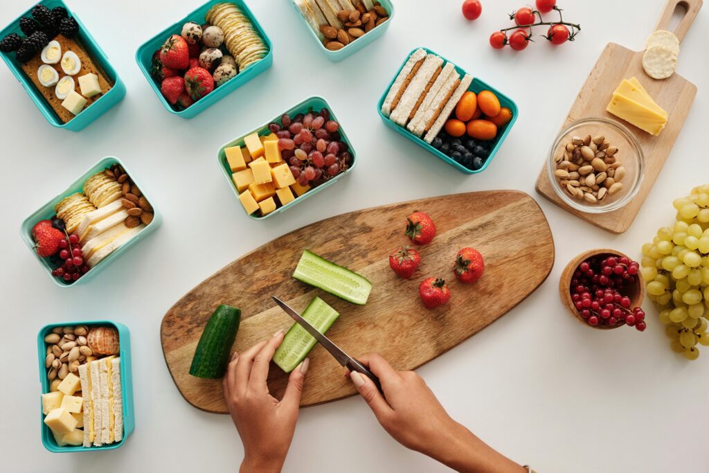An overhead shot shows a person's hands slicing a cucumber on a wooden cutting board, surrounded by several teal bento boxes filled with various snacks. The containers include finger sandwiches, cheese cubes, grapes, strawberries, blackberries, hard-boiled eggs, almonds, pistachios, and crackers. Additional fresh produce like cherry tomatoes, red currants, and a bunch of green grapes are scattered across the clean white surface.
