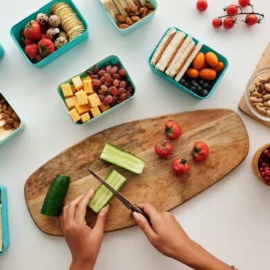 An overhead shot shows a person's hands slicing a cucumber on a wooden cutting board, surrounded by several teal bento boxes filled with various snacks. The containers include finger sandwiches, cheese cubes, grapes, strawberries, blackberries, hard-boiled eggs, almonds, pistachios, and crackers. Additional fresh produce like cherry tomatoes, red currants, and a bunch of green grapes are scattered across the clean white surface.