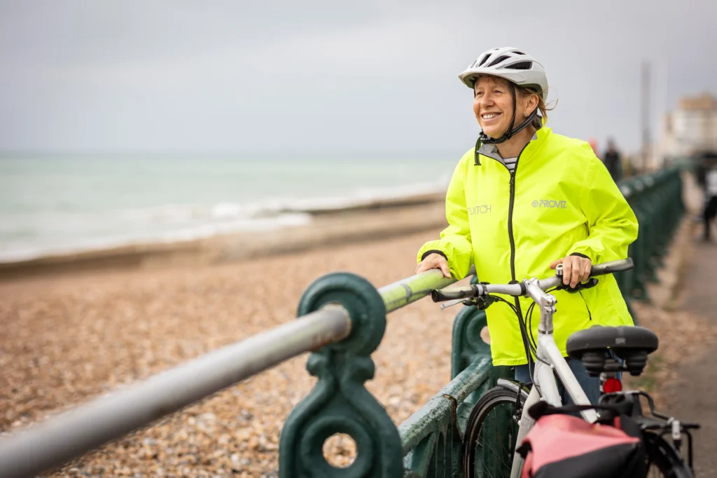 A smiling woman wearing a white helmet and a bright neon yellow high-visibility jacket stands with her bicycle next to a green railing overlooking a pebble beach and the ocean.