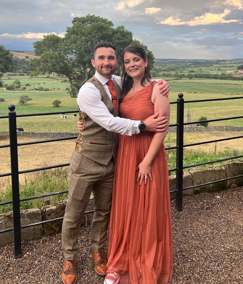 A full-length photograph of Amy and Ben posing closely together outdoors against a scenic countryside backdrop. Amy, on the right, wears a long, sleeveless burnt-orange dress and pink Converse. Ben, on the left, wears a tan plaid three-piece suit with an orange tie and brown brogues, with his arm around Amy’s shoulder. They are standing on a gravel terrace behind a black metal railing. The background features rolling green hills, scattered trees, and a dramatic, cloudy sky with hints of sunlight.
