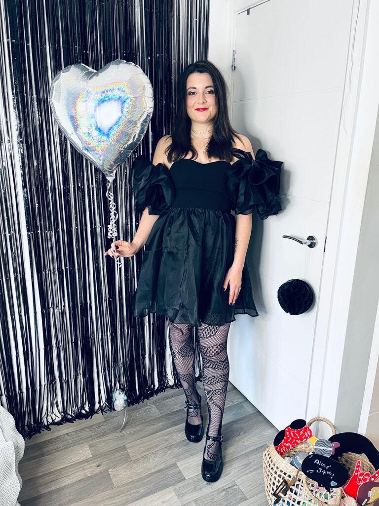 A full-length photo of Amy posing for her birthday celebration. She is wearing a black off-the-shoulder mini dress with voluminous ruffled sleeves, patterned fishnet tights, and black Mary Jane shoes. She has shoulder-length dark hair and is smiling while holding a silver heart-shaped balloon. She is standing in a room with a black tinsel backdrop to her left and a white door to her right. In the bottom right corner, there is a wicker basket filled with party props, including a sign that reads "Aims' 34th!"