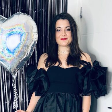 Amy posing for a photo while holding a silver, heart-shaped holographic balloon. She is wearing a black off-the-shoulder dress with large ruffled sleeves and red lipstick. She is standing in front of a black tinsel backdrop and a white door.