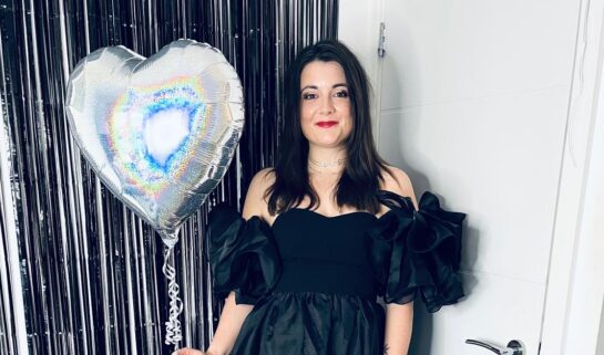 Amy posing for a photo while holding a silver, heart-shaped holographic balloon. She is wearing a black off-the-shoulder dress with large ruffled sleeves and red lipstick. She is standing in front of a black tinsel backdrop and a white door.