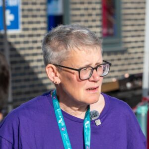 Ceri has short grey hair and glasses, and wears a purple t-shirt and a teal lanyard.