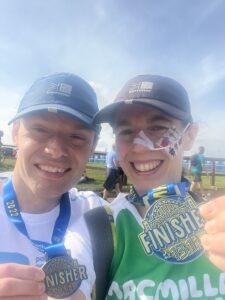 A selfie of Paul and Danielle smiling after completing the Great North Run. Both are wearing running caps and proudly holding up their "Finisher 13.1 Miles" medals. Danielle is wearing a green Macmillan Cancer Support shirt and has a feeding tube secured to her cheek with medical tape.