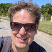 David is enjoying a sunny bike ride and wears black sunglasses. The sky is blue and he has light brown hair.