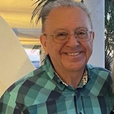 Leslie wears a teal and navy checkered short-sleeved shirt, smiling warmly at the camera.