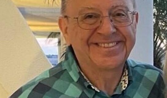 Leslie wears a teal and navy checkered short-sleeved shirt, smiling warmly at the camera.