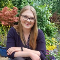 Megan is smiling and has long blonde hair. She wears glasses and sits in a lush, green garden. She is wearing a dark purple cardigan over a floral top, with her arms crossed in front of her. The background features a variety of vibrant plants.