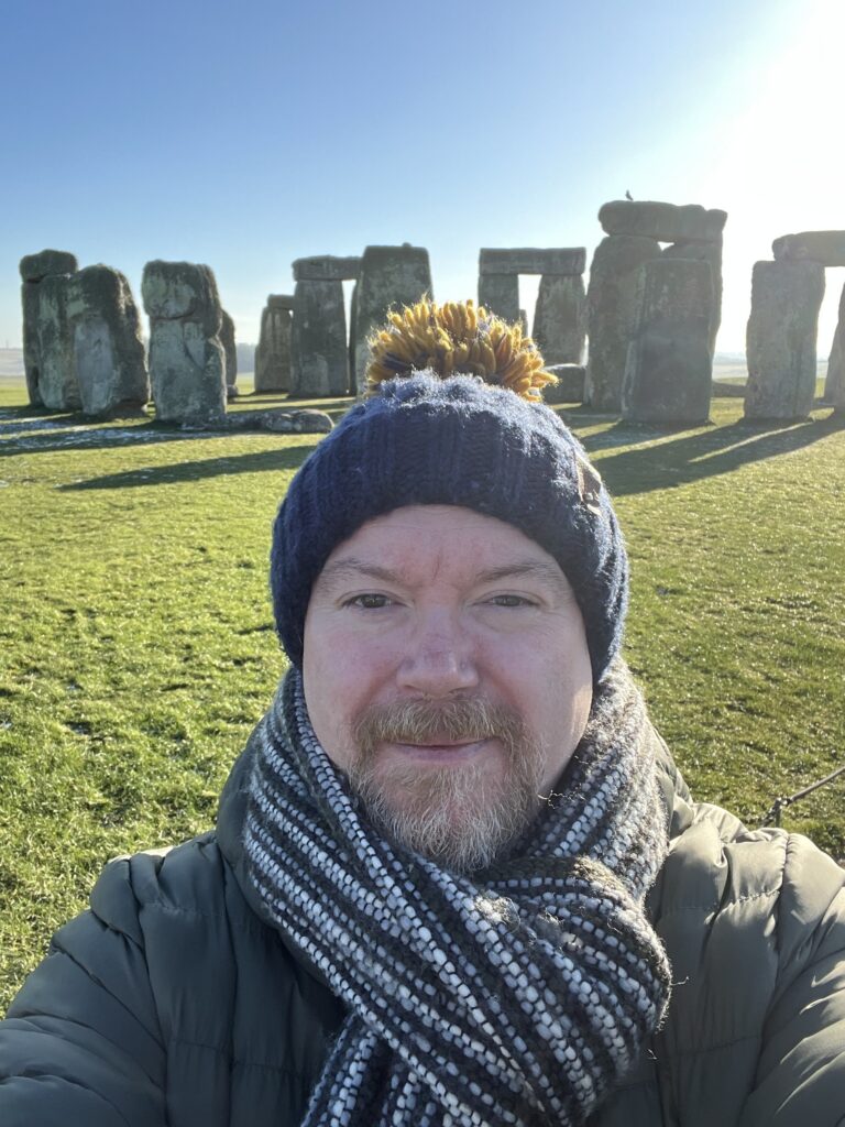 A selfie of Peter, a man with a light-coloured beard and moustache, wearing a blue knit beanie with a yellow pom-pom and a thick, patterned grey scarf. He is standing outdoors in a grassy field with the ancient stone circle of Stonehenge visible in the background under a bright, clear sky.