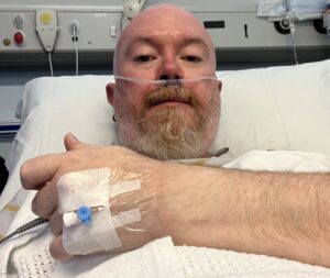A close-up selfie of Peter lying in a hospital bed. He has a nasal cannula for oxygen and a medical IV port taped to the back of his hand, which is resting on a white honeycomb-patterned blanket. The hospital headboard with various medical outlets and buttons is visible behind him.