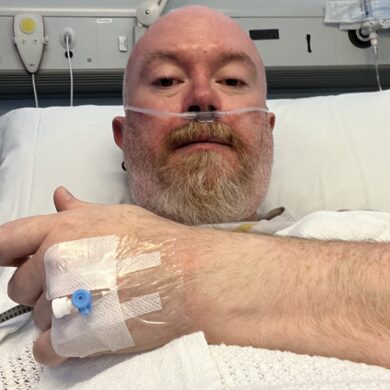 A close-up selfie of Peter lying in a hospital bed. He has a nasal cannula for oxygen and a medical IV port taped to the back of his hand, which is resting on a white honeycomb-patterned blanket. The hospital headboard with various medical outlets and buttons is visible behind him.