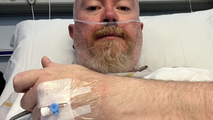 A close-up selfie of Peter lying in a hospital bed. He has a nasal cannula for oxygen and a medical IV port taped to the back of his hand, which is resting on a white honeycomb-patterned blanket. The hospital headboard with various medical outlets and buttons is visible behind him.