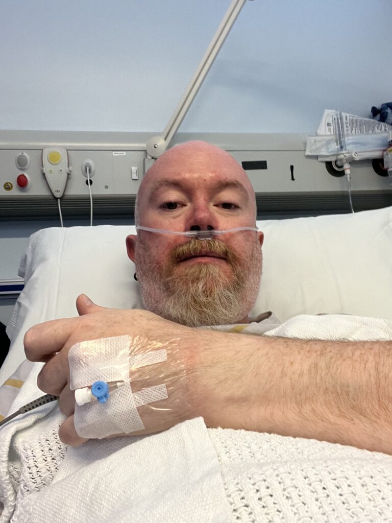 A close-up selfie of Peter lying in a hospital bed. He has a nasal cannula for oxygen and a medical IV port taped to the back of his hand, which is resting on a white honeycomb-patterned blanket. The hospital headboard with various medical outlets and buttons is visible behind him.