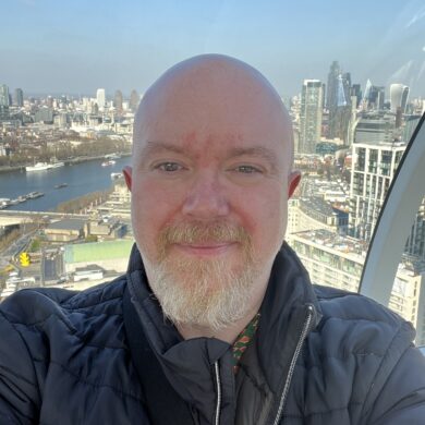 A selfie of Peter taken from a high vantage point, likely inside a glass-walled observation wheel like the London Eye. He is smiling slightly, wearing a black puffer jacket. Behind him, a panoramic view of the London skyline and the River Thames stretches out under a blue sky.