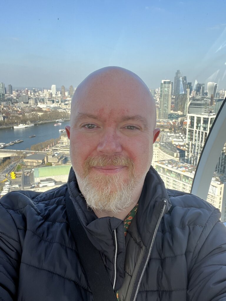A selfie of Peter taken from a high vantage point, likely inside a glass-walled observation wheel like the London Eye. He is smiling slightly, wearing a black puffer jacket. Behind him, a panoramic view of the London skyline and the River Thames stretches out under a blue sky.