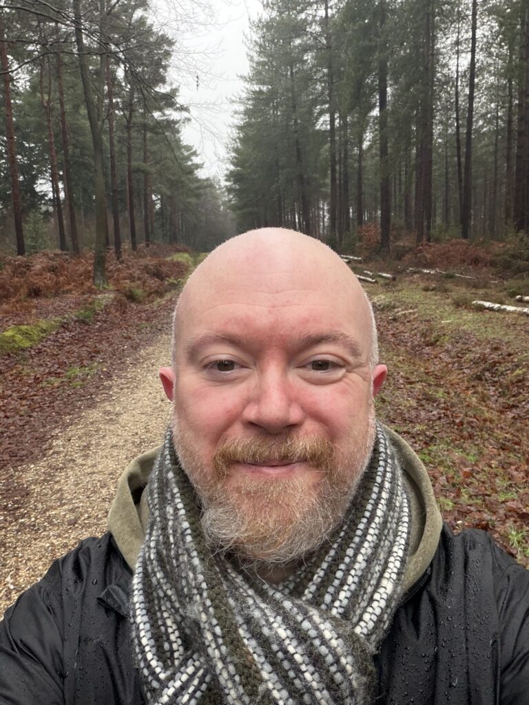 A selfie of Peter standing on a dirt path in the middle of a dense forest. He is wearing a black raincoat, a green hoodie, and a striped wool scarf. The tall, thin trees and fallen leaves suggest a cool, overcast day in a woodland setting.