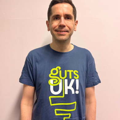 Richard is wearing a navy Guts UK t-shirt and stands against a cream wall. He is smiling at the camera and has short, spiky, black hair.