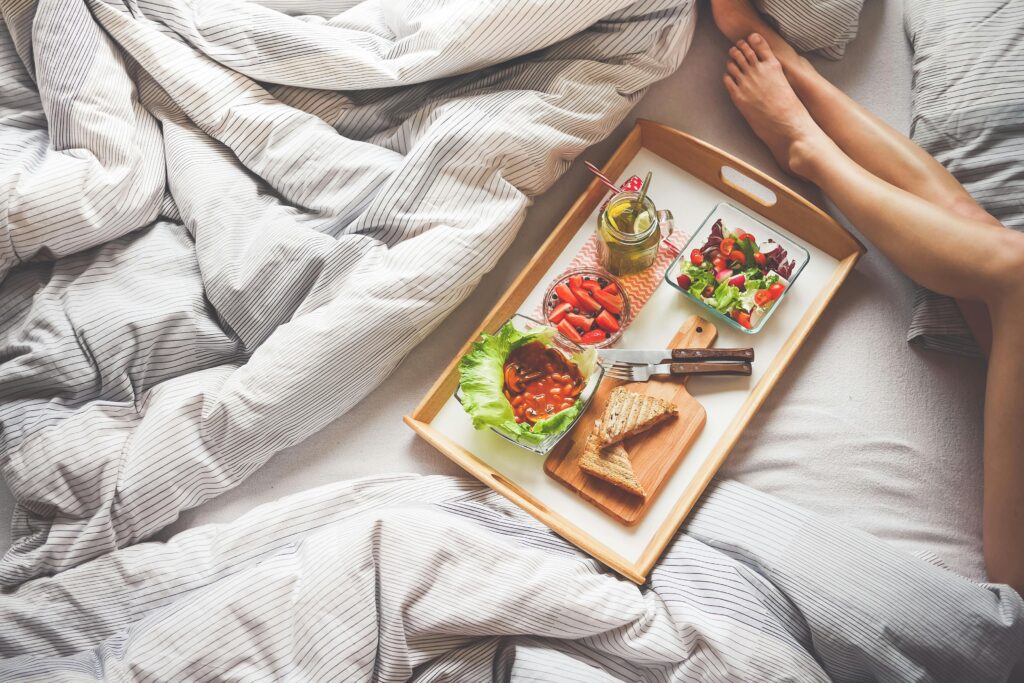 A high-angle view of a wooden tray with breakfast in bed on striped white and grey linens. The tray holds toasted sandwiches, baked beans, a garden salad, sliced strawberries, and a green drink with lemon. A person's legs are visible resting on the bed beside the meal.