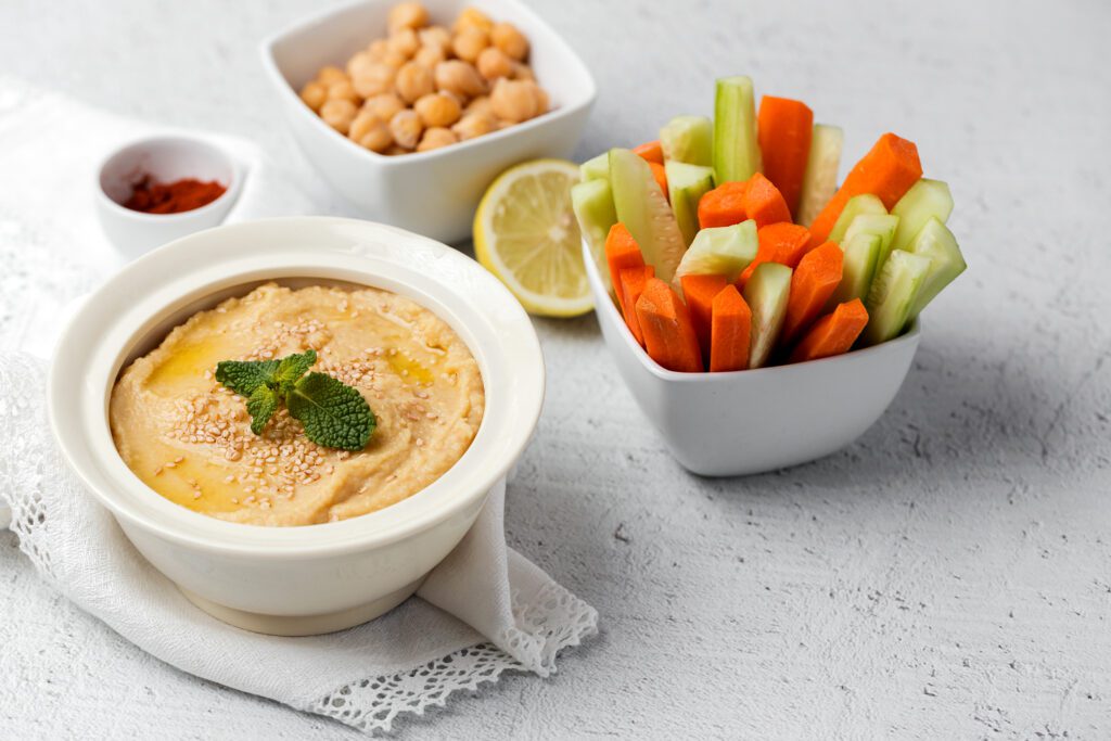  A small white bowl of smooth hummus topped with sesame seeds and a sprig of mint, served on a white cloth alongside a bowl of carrot and cucumber sticks, a bowl of whole chickpeas, and a lemon half.