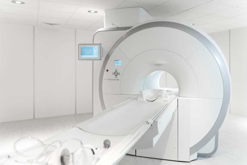 A wide shot of a sleek, white MRI machine in a brightly lit, sterile room. The large, circular scanner features a central bore and a patient table extended outward. A small monitor is mounted on the side of the machine, and the minimalist background consists of white paneled walls and a clean grey floor.