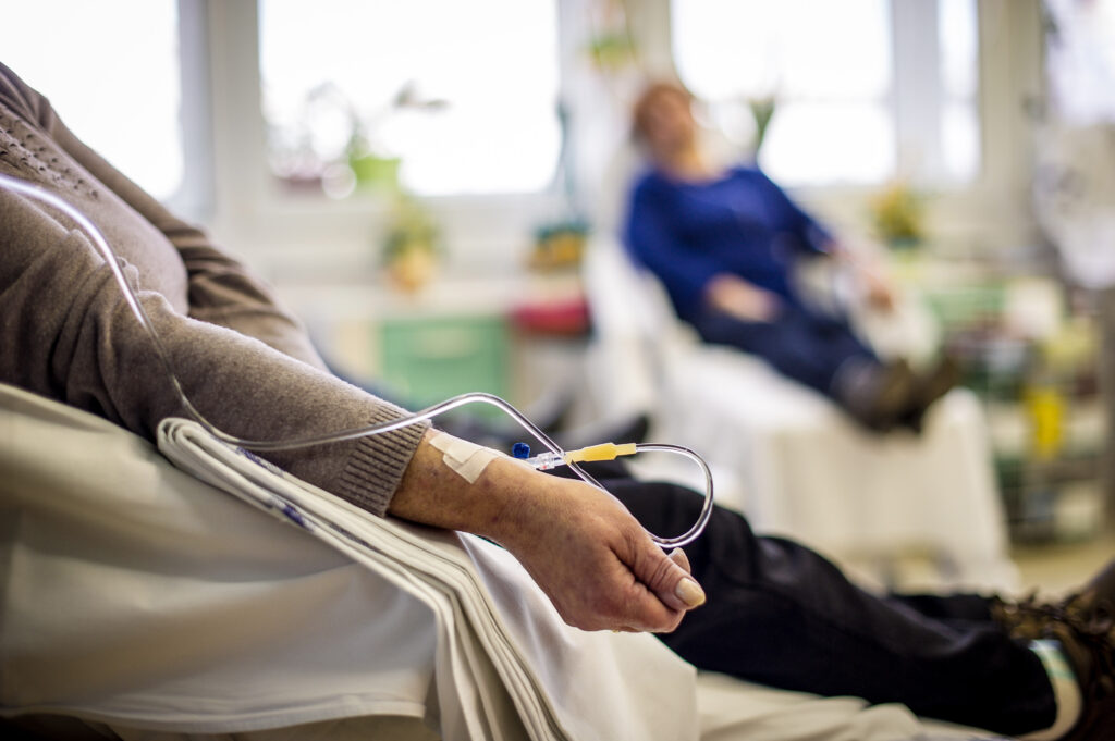 A close-up of a patient's hand resting on a white hospital bed with an IV drip line inserted into their arm. Another person is visible in the blurred background, also receiving treatment in a bright clinic setting.