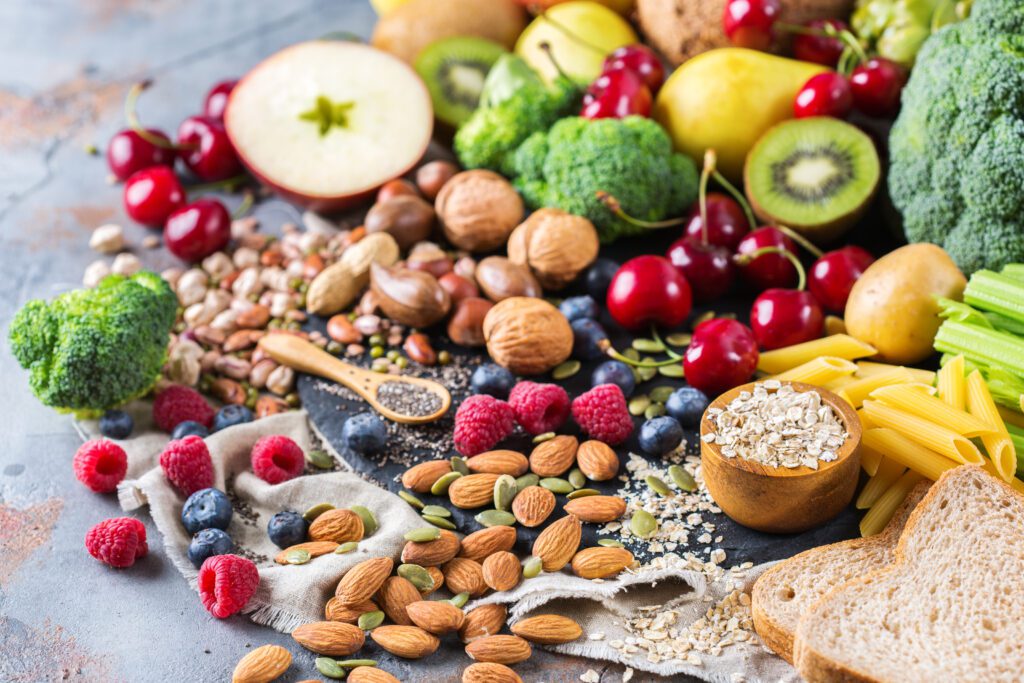 A vibrant, top-down arrangement of diverse whole foods spread across a rustic gray surface. In the foreground, a cluster of whole almonds and green pumpkin seeds sits on a linen cloth next to scattered raspberries and blueberries. To the right, a small wooden bowl holds rolled oats next to slices of whole-grain bread and a pile of penne pasta. The center features various nuts, including walnuts and hazelnuts, and a small wooden spoon filled with chia seeds. The background is filled with fresh produce, including broccoli florets, cherries, kiwi halves, a sliced apple, pears, and stalks of celery. The composition emphasizes a balanced, plant-based diet rich in fiber and nutrients.