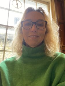 Alex is wearing a bright green jumper and dark-rimmed glasses. She has blonde curly hair and is smiling. Behind her, is a large window with light pouring in.