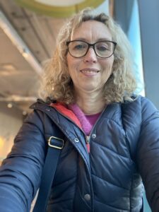 Alex is smiling at the camera. She is wearing a navy blue quilted puffer jacket. She has blonde curly hair and wears dark-rimmed glasses.