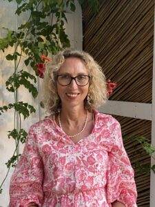 Alex has blonde curly hair and dark-rimmed glasses is smiling at the camera. She is wearing a pink and white floral patterned shirt dress and a pearl necklace. She stands in front of a climbing plant with green leaves and orange bell-shaped flowers.