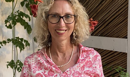 Alex has blonde curly hair and dark-rimmed glasses is smiling at the camera. She is wearing a pink and white floral patterned shirt dress and a pearl necklace. She stands in front of a climbing plant with green leaves and orange bell-shaped flowers.