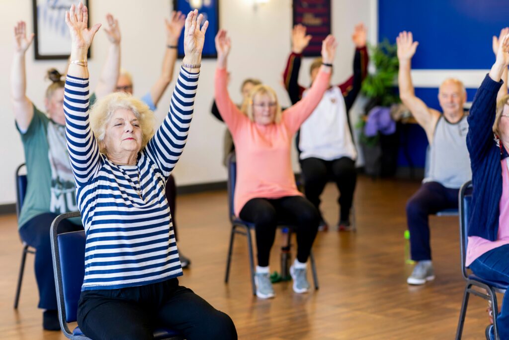 A group of older adults participate in a chair-based exercise class in a bright room, all raising their arms overhead while seated.
